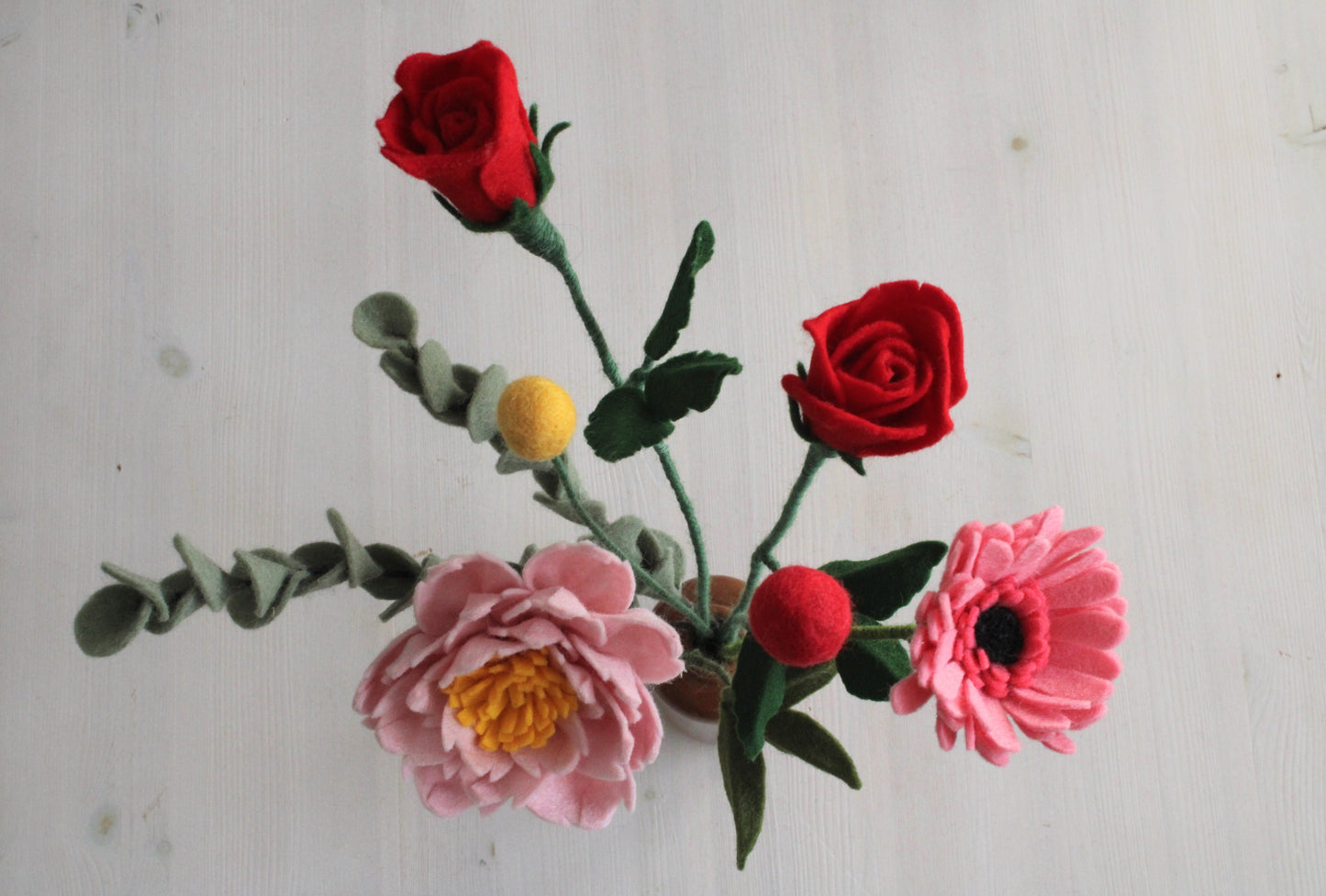 Handgemaakt viltbloemenboeket met rode rozen, gele en rode craspedia, roze pioenroos, roze gerbera en twee eucaluptus takken in een bruine vaas op een witte ondergrond