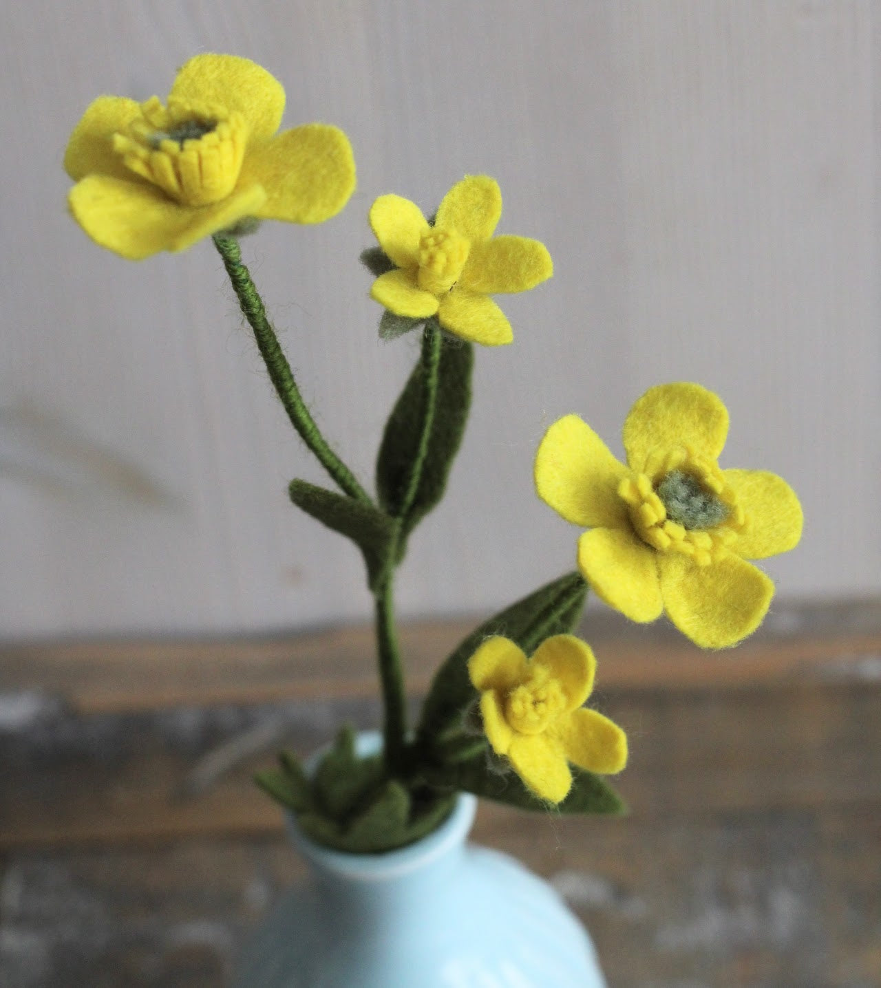 Boterbloem van geel vilt, handgemaakt en ideaal voor een duurzaam bloemenboeketje. in een blauw vaasje op een rustieke houten ondergrond