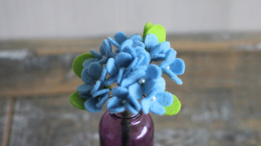 Close-up van lichtblauwe mini hortensia met groene blaadjesgemaakt van vilt in een paars vaasje met een houten achtergrond