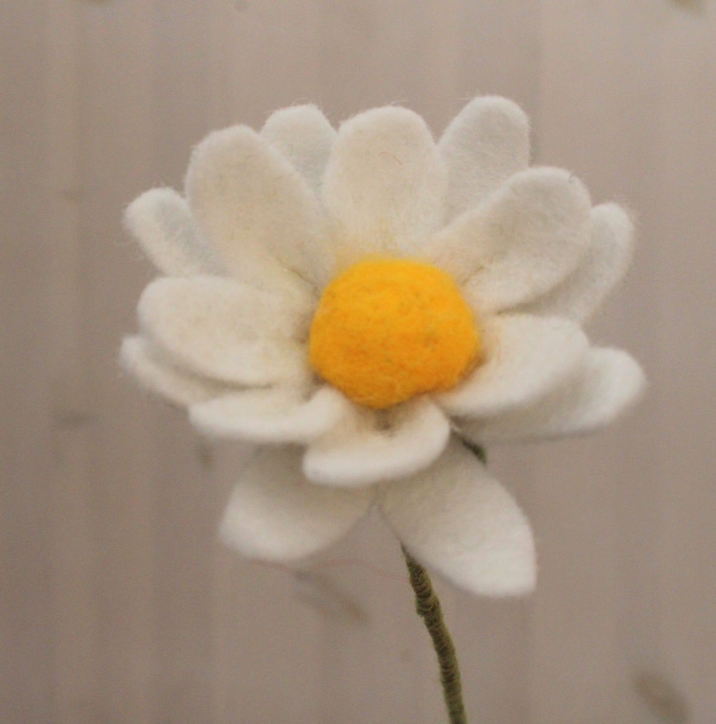 Handgemaakte vilten margriet met witte bloemblaadjes en een geel hart, op een lange steel bekleed met groene wol. Geplaatst in een bruine keramieken vaas met een lichte achtergrond. Perfect als duurzame decoratie of als onderdeel van een viltbloemenboeket