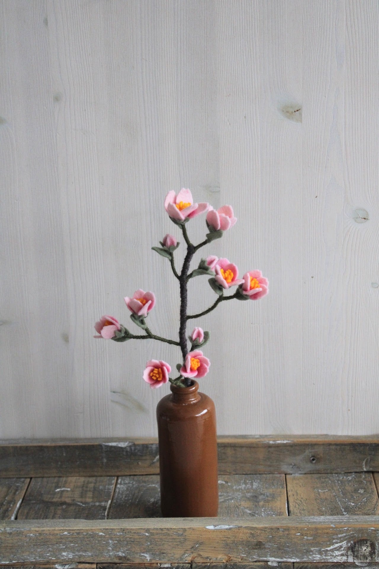 Handgemaakte vilten kersenbloesemtak met roze bloemetjes en een geel hartje, op een korte steel bekleed met bruine wol. Geplaatst in een bruine vaas op een rustieke houten ondergrond. Perfect als duurzame decoratie of als onderdeel van een viltbloemenboeket.