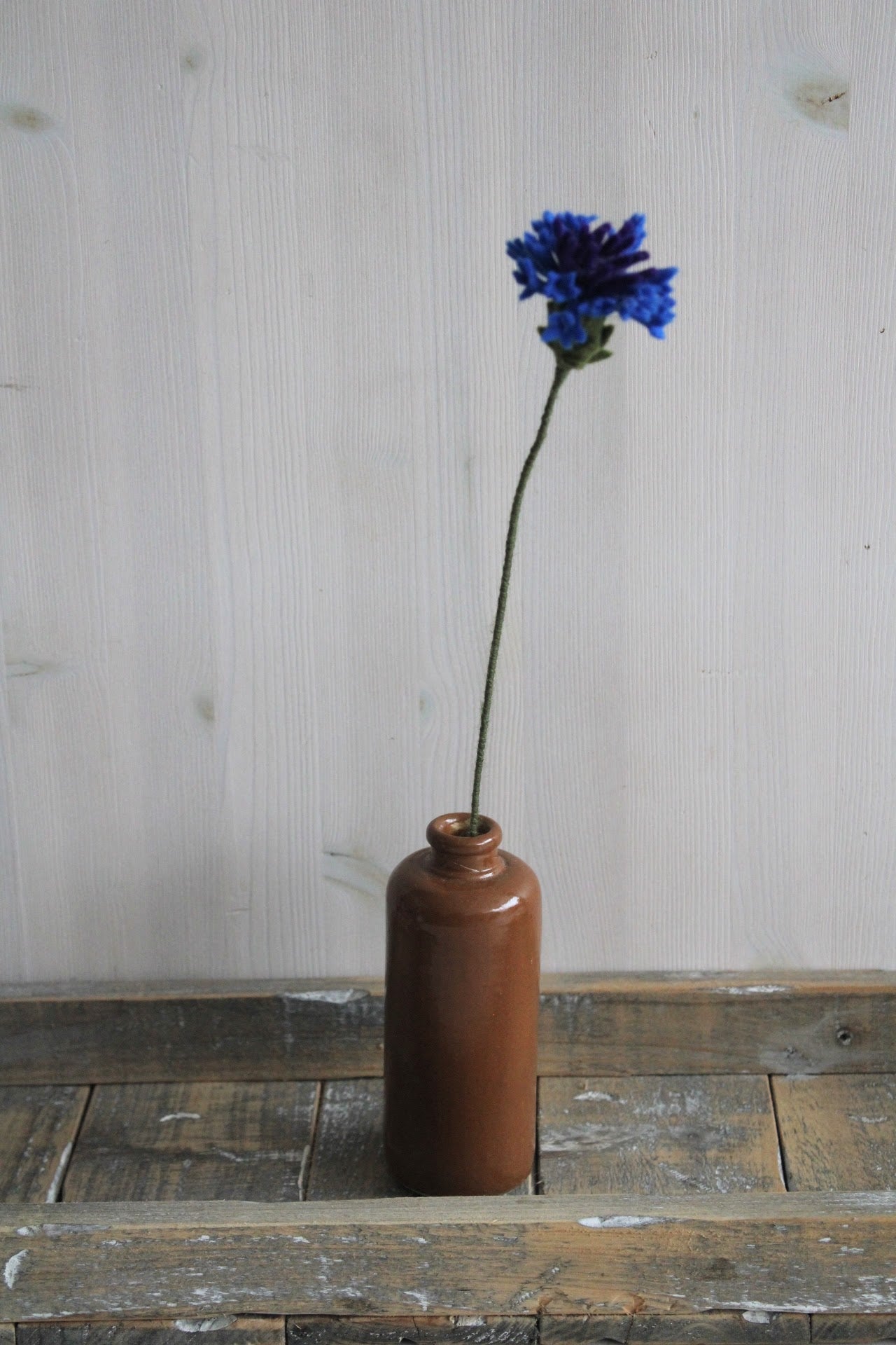 Handgemaakte vilten korenbloem met blauwe bloemblaadjes, op een lange steel bekleed met groene wol. Geplaatst in een bruine keramieken vaas op een rustieke houten ondergrond. Perfect als duurzame decoratie of als onderdeel van een viltbloemenboeket