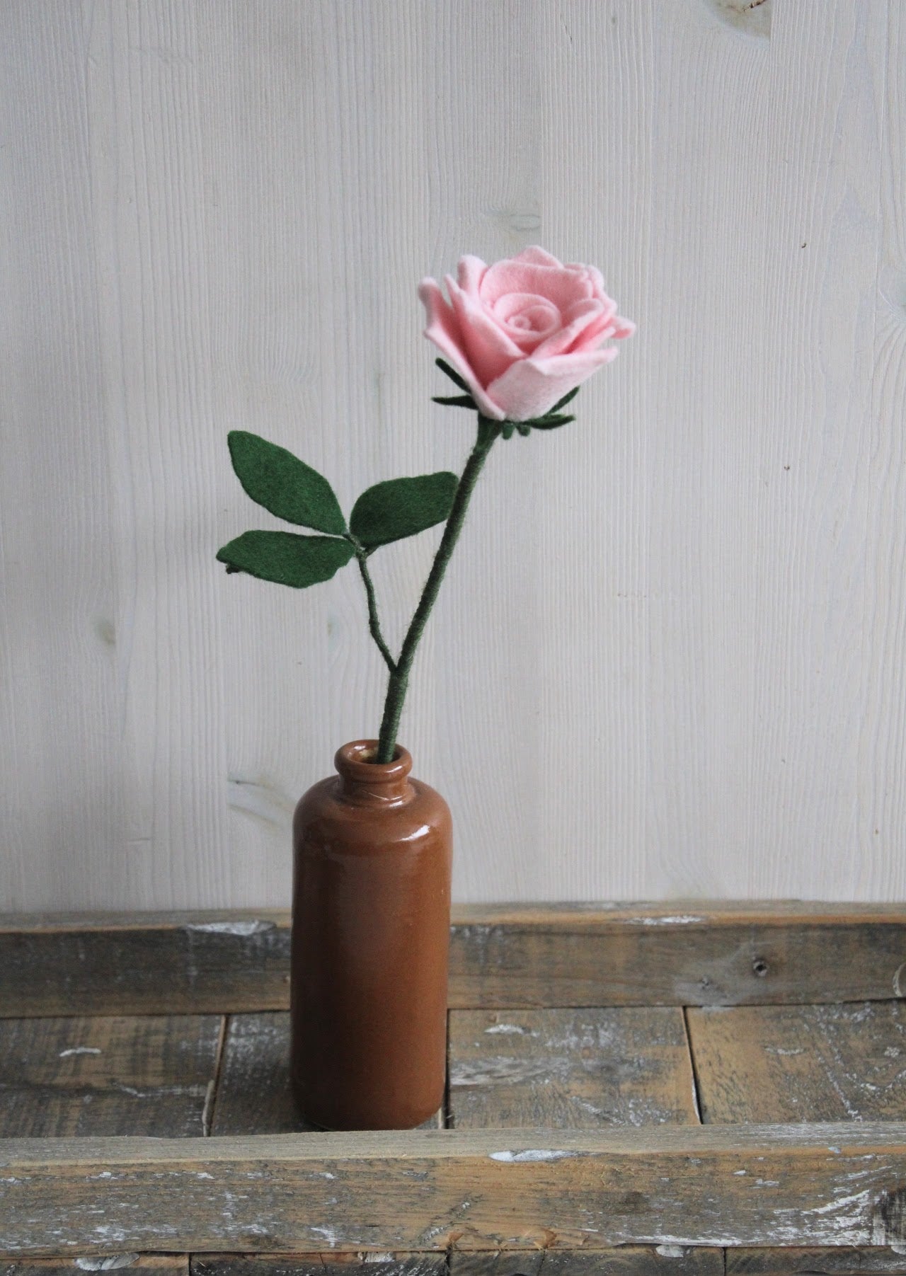 Handgemaakte vilten roze roos op lange steel bekleed met groene wol en blad. Geplaatst in een bruine keramieken vaas op een rustieke houten ondergrond. Perfect als duurzame decoratie of in een blijvend bloeiend viltbloemenboeket