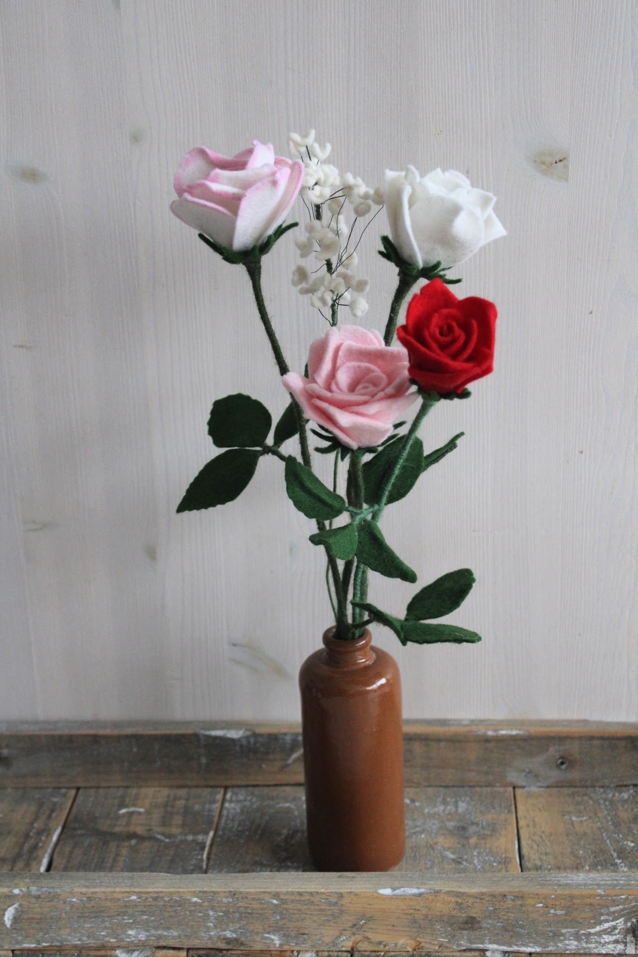 Handgemaakt boeket van viltbloemen in een bruine vaas, met roze, rode en witte rozen en fijn wit gipskruid tegen een witte achtergrond en houten ondergrond