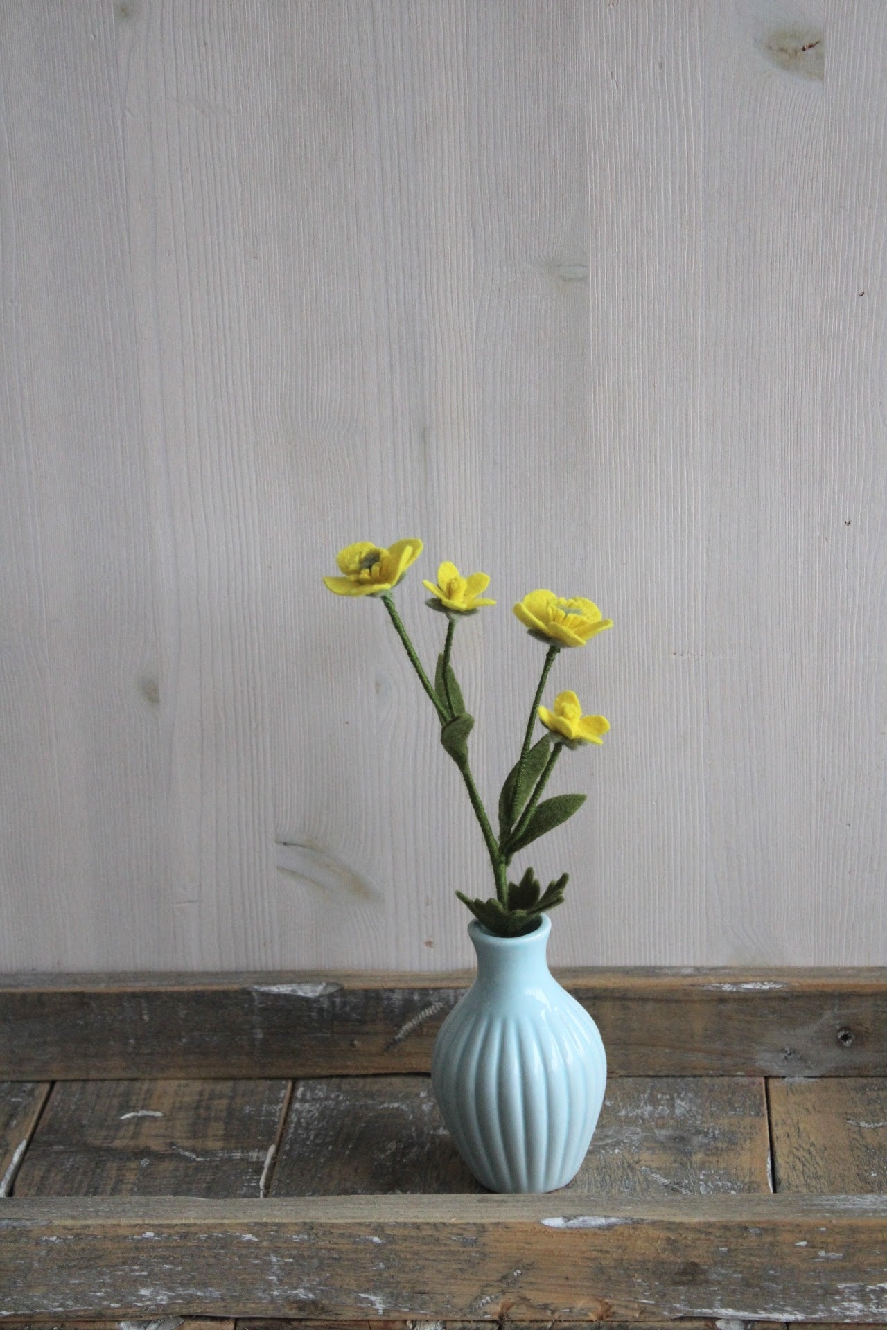 Handgemaakte boterbloem van vilt met 4 bloemetjes en blaadjes in blauw vaasje op een rustieke houten ondergrond