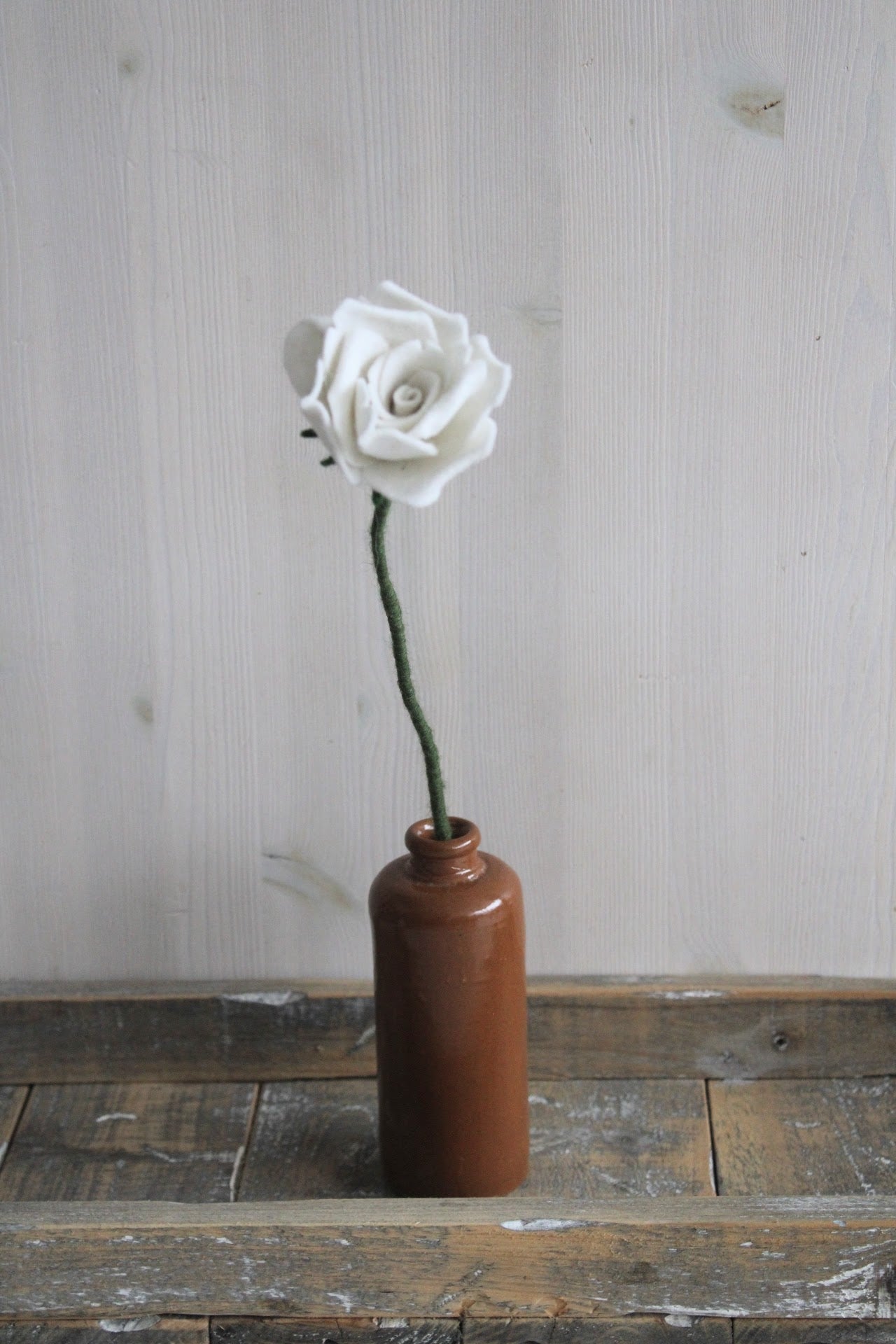 Handgemaakte vilten witte roos op lange steel bekleed met groene wol. Geplaatst in een bruine keramieken vaas op een rustieke houten ondergrond. Perfect als duurzame decoratie of in een blijvend bloeiend viltbloemenboeket