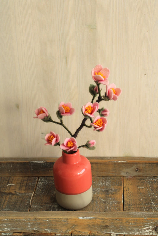 Keramisch vaasje met een moderne tweekleurige afwerking: een matte beige onderkant en een glanzende koraalrode bovenkant met een tak kersenbloesem op een houten rustieke plank