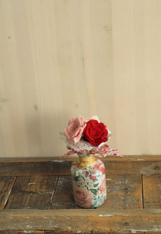 Biedermeiertje van drie roosjes afgewerkt met kant en een gebloemde strik in een gebloemd fles vaasje op een houten ondergrond