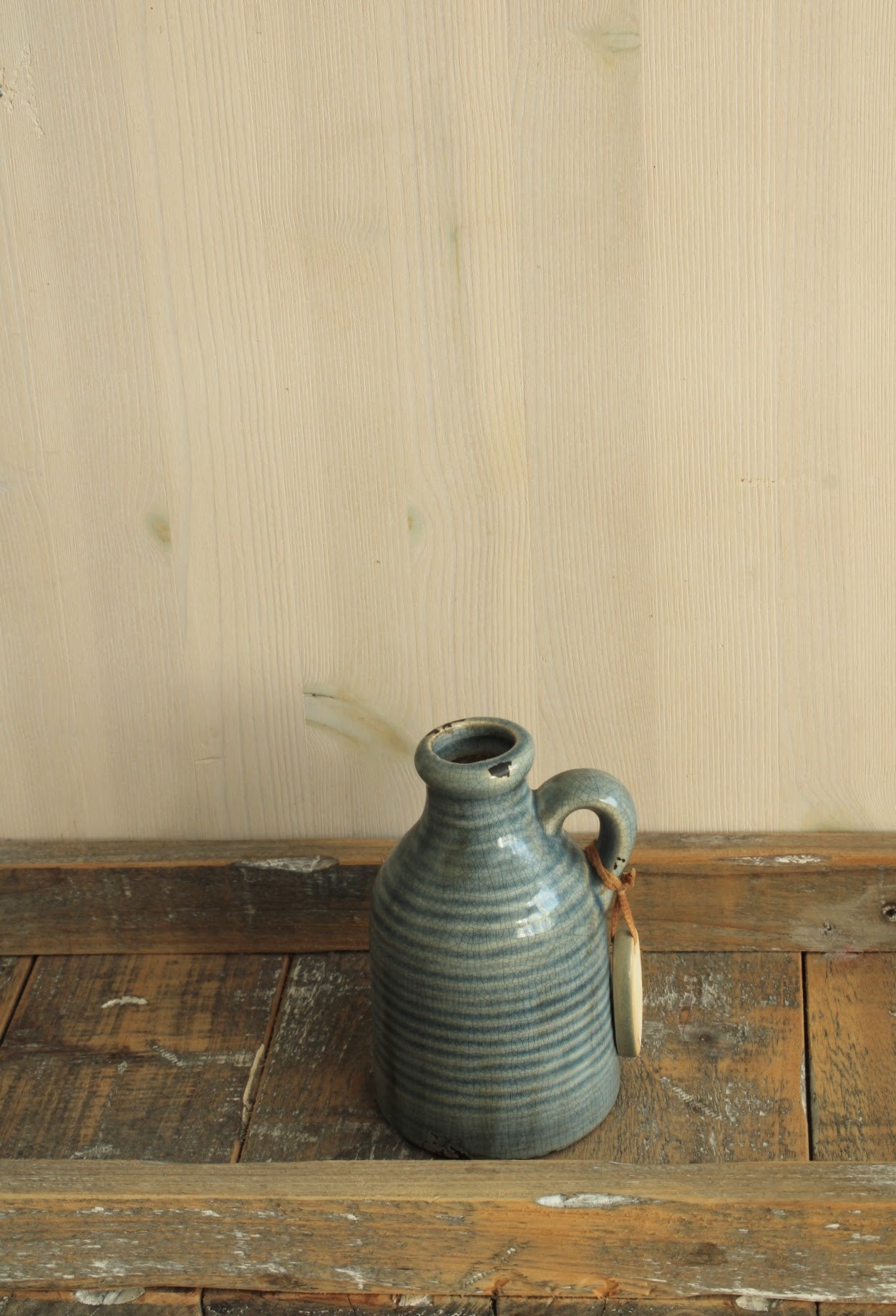 Rustiek keramisch kruikvaasje met blauwgrijze strepen, een handvat en een decoratief label op een houten ondergrond