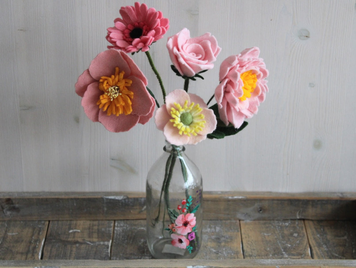 Vilten boeket met verschillende roze bloemen, gerbera, pioenroos, roos, twee anemonen in een glazen vaas op en houten ondergrond