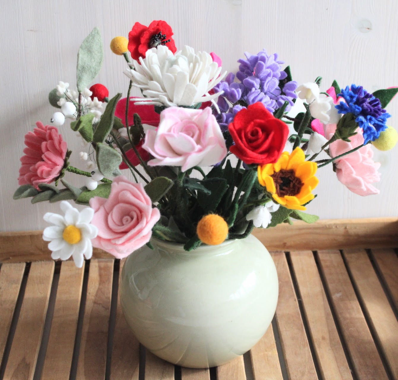 Uitbundig boeket van handgemaakte vilten bloemen in een bolvormige vaas: rozen in rood, roze en wit; chrysant; klaprozen; lelietjes-van-dalen; gerbera; zonnebloem; korenbloem; vergeet-mij-nietjes en diverse andere bloemen en groen.