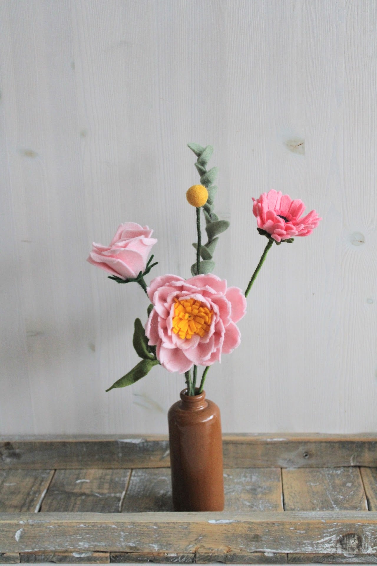 Vilten boeket met verschillende roze bloemen, gerbera, pioenroos, roos, eucalyptus tak en gele craspedia in een bruine vaas op en houten ondergrond