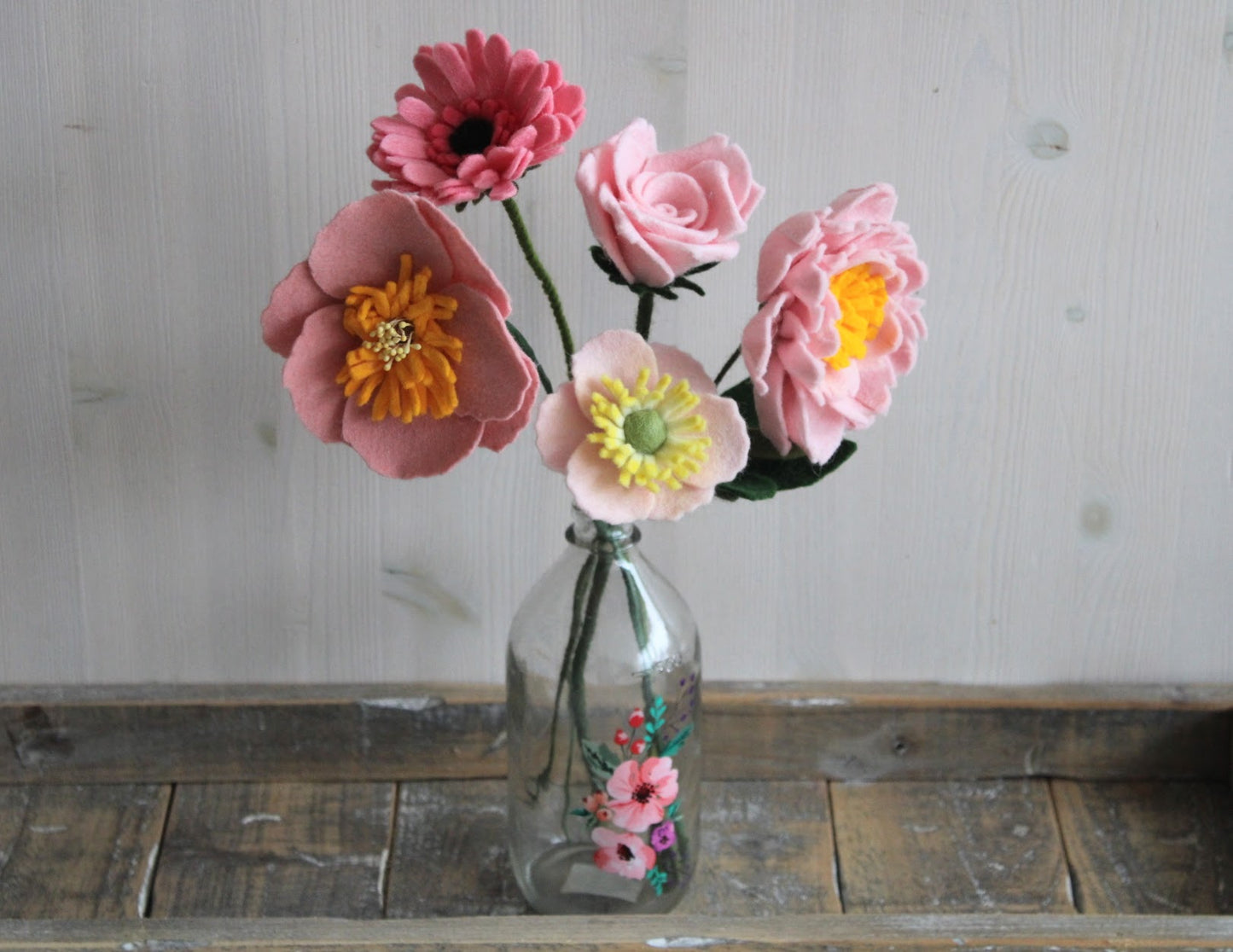 Vilten boeket met verschillende roze bloemen, gerbera, pioenroos, roos, twee anemonen in een glazen vaas op en houten ondergrond