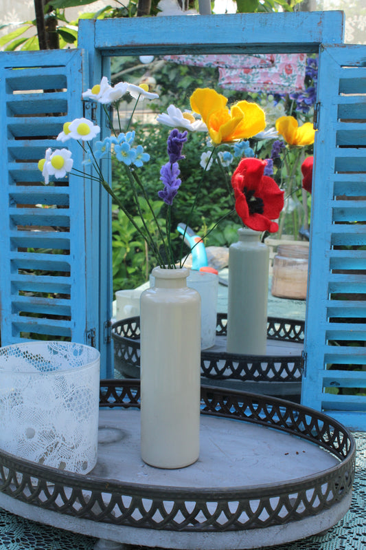 Handgemaakt boeket viltbloemen in een witte vaas, met madeliefjes, lavendel, vergeet-me-nietjes, gele tulpen en rode klaproos, gepresenteerd voor een blauwe spiegel met tuin op de achtergrond