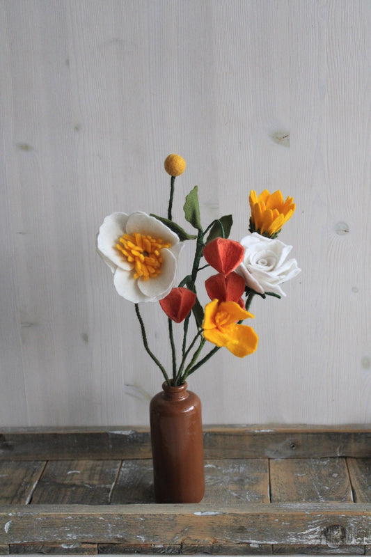 Zomers handgemaakt viltbloemen boeket met oranje lampionnetje, geel slaapmutsje, zonnebloem, witte roos, witte anemoon en gele craspedia in een bruine vaas op een rustikeke houten