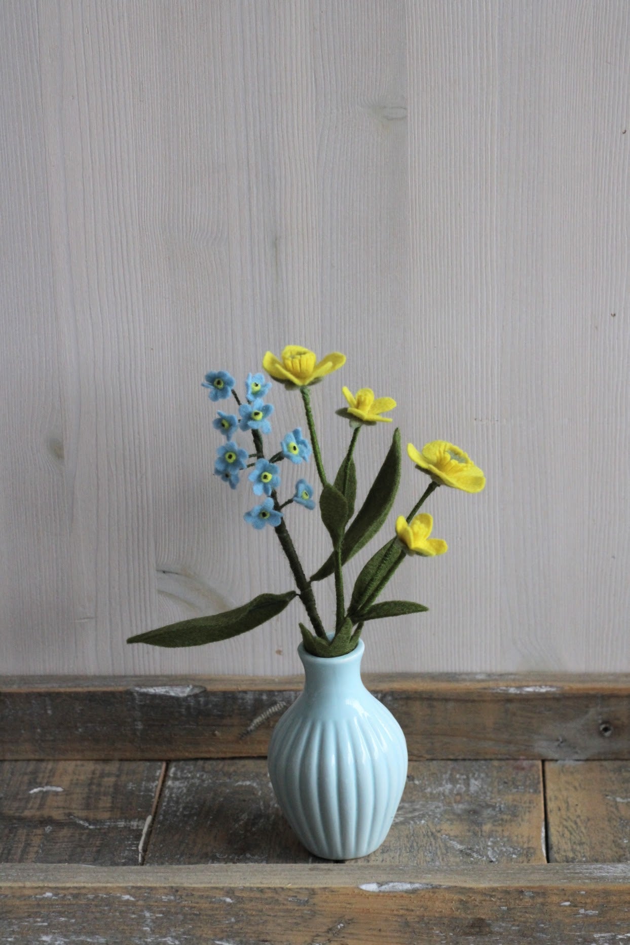 Gele boterbloem en een blauw vergeetmenietje van vilt in een blauw vaasje op een houten plank, handgemaakt en ideaal voor een duurzaam bloemenboeketje.
