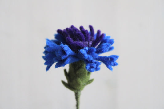 Handgemaakte vilten korenbloem met blauwe bloemblaadjes, op een lange steel bekleed met groene wol, met een lichte achtergrond. Perfect als duurzame decoratie of als onderdeel van een viltbloemenboeket