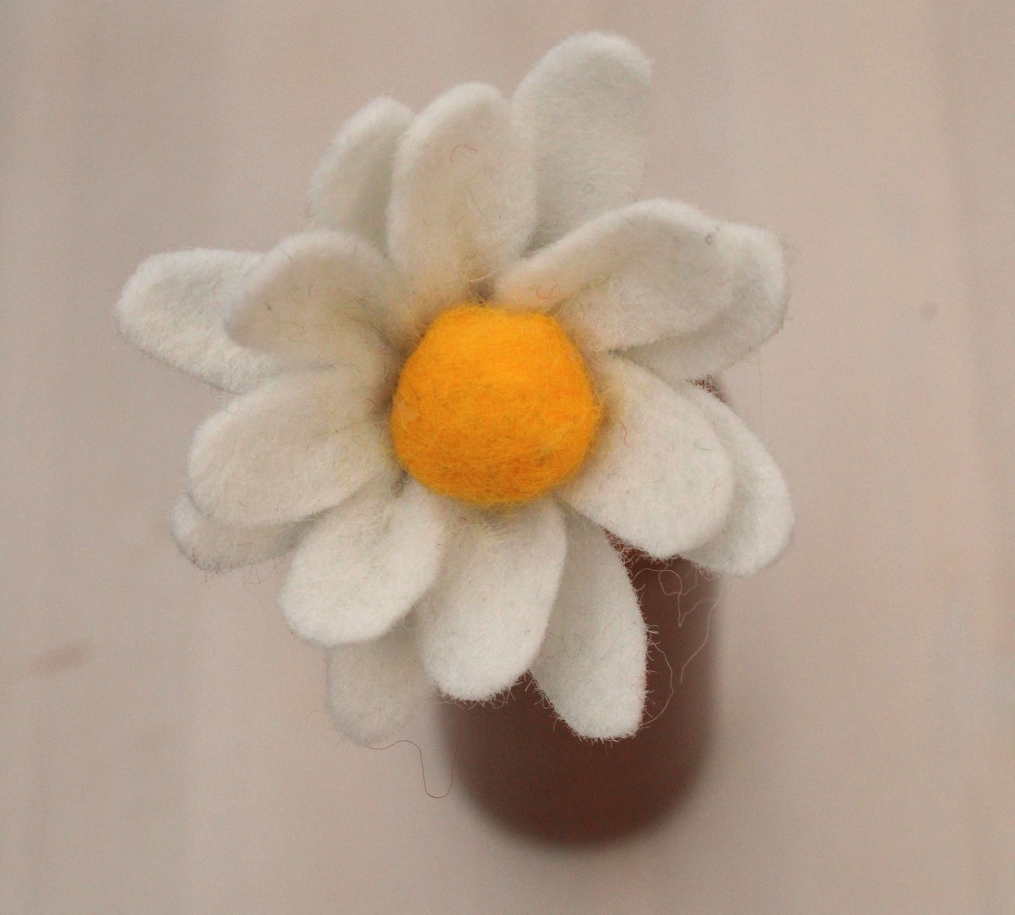 Handgemaakte vilten margriet met witte bloemblaadjes en een geel hart, op een lange steel bekleed met groene wol. Geplaatst in een bruine keramieken vaas op een witte ondergrond. Perfect als duurzame decoratie of als onderdeel van een viltbloemenboeket
