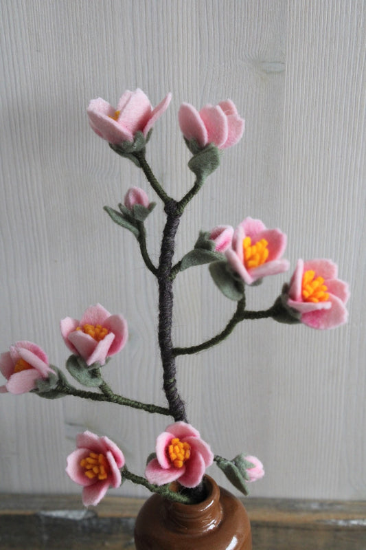 Handgemaakte kersenbloesemtak van vilt met zachte roze bloemetjes en groene blaadjes, geplaatst in een bruine keramieken vaas op een rustieke houten ondergrond. Een tijdloze en duurzame decoratie, perfect voor een lentegevoel in huis