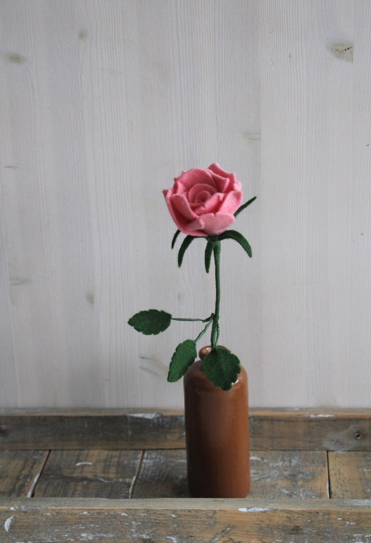 Handgemaakte vilten roze roos op lange steel bekleed met groene wol en blad. Geplaatst in een bruine keramieken vaas op een rustieke houten ondergrond. Perfect als duurzame decoratie of in een blijvend bloeiend viltbloemenboeket