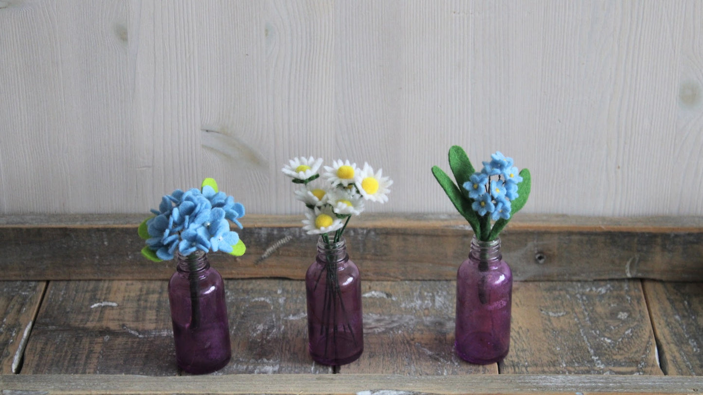 3 boeketjes met blauwe hortensia/madeliefjes/ blauw vergeet-me-nietje in een paars vaasje op een houten ondergrond en lichte achtergrond