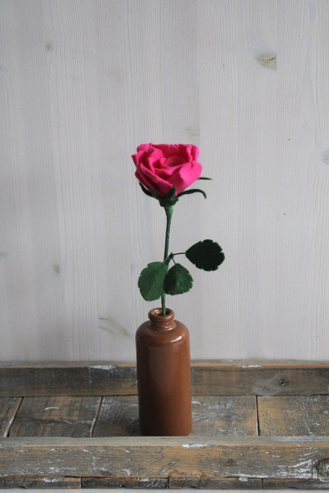 Handgemaakte vilten felroze roos op lange steel bekleed met groene wol en blad. Geplaatst in een bruine keramieken vaas op een rustieke houten ondergrond. Perfect als duurzame decoratie of in een blijvend bloeiend viltbloemenboeket