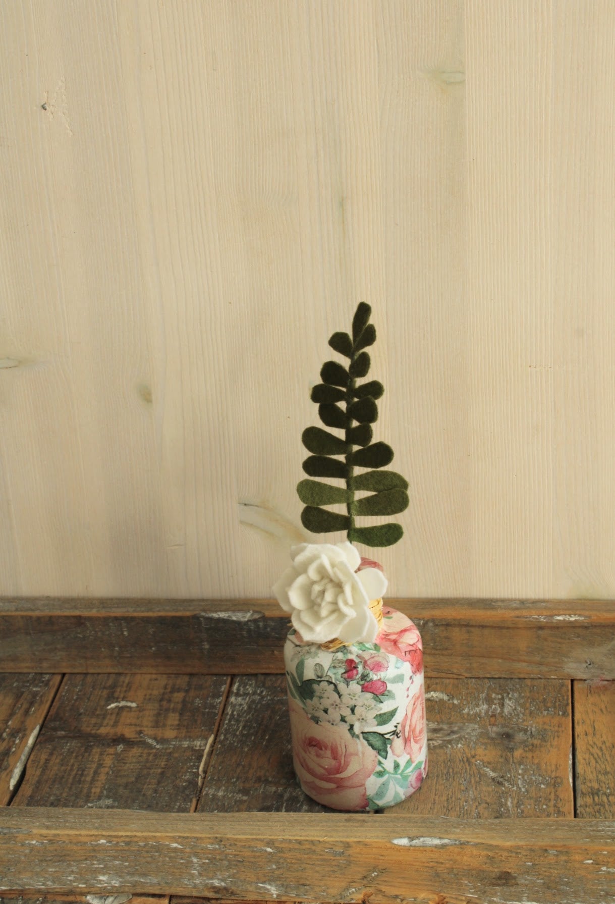 gebloemd fles vaasje met 1 takje bladgroen en witte bloem op een houten rustieke plank