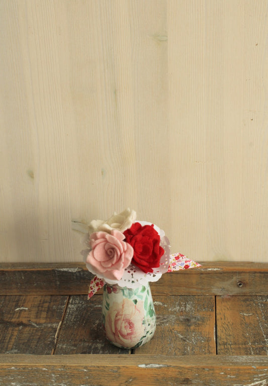 Gebloemd vaasje met biedermeiertje van drie roosjes in de kleur wit, roze en rood met kant afgewerkt en een gebloemde strik op een houten ondergrond