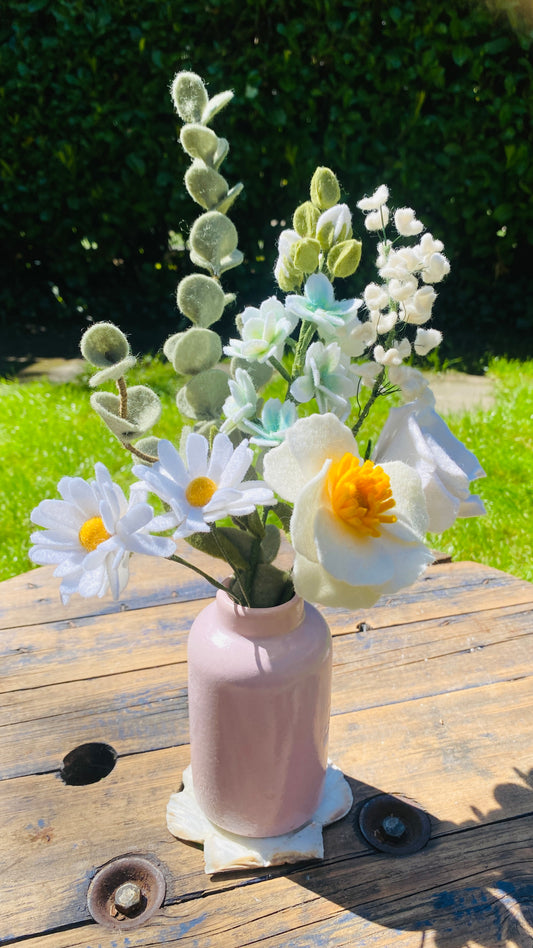 Gemengd wit boeket viltbloemen met margrieten, eucalyptus, gipskruid, violier, anemoon en roos in een roze vaasje op een houten tafel en gras op de achtergrond