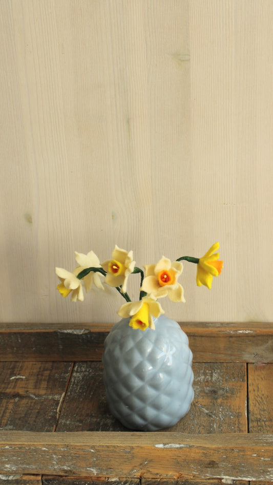 Grijs keramisch vaasje met ruitpatroon, met schattige narcisjes, geplaatst op een rustiek houten dienblad tegen een lichte houten achtergrond.