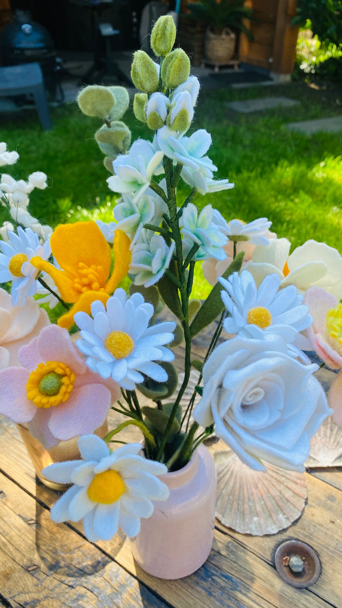 Gemengd wit boeket viltbloemen met margrieten,eucalyptus, gipskruid, violier, anemoon, geel slaapmutsje en witte roos in een roze vaasje op een houten tafel met grote schelpen en gras op de achtergrond