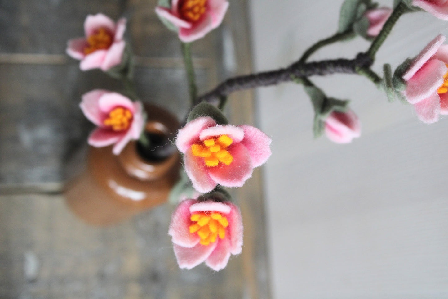 Handgemaakte kersenbloesemtak van vilt met zachte roze bloemetjes en groene blaadjes, geplaatst in een bruine keramieken vaas op een rustieke houten ondergrond. Een tijdloze en duurzame decoratie, perfect voor een lentegevoel in huis.