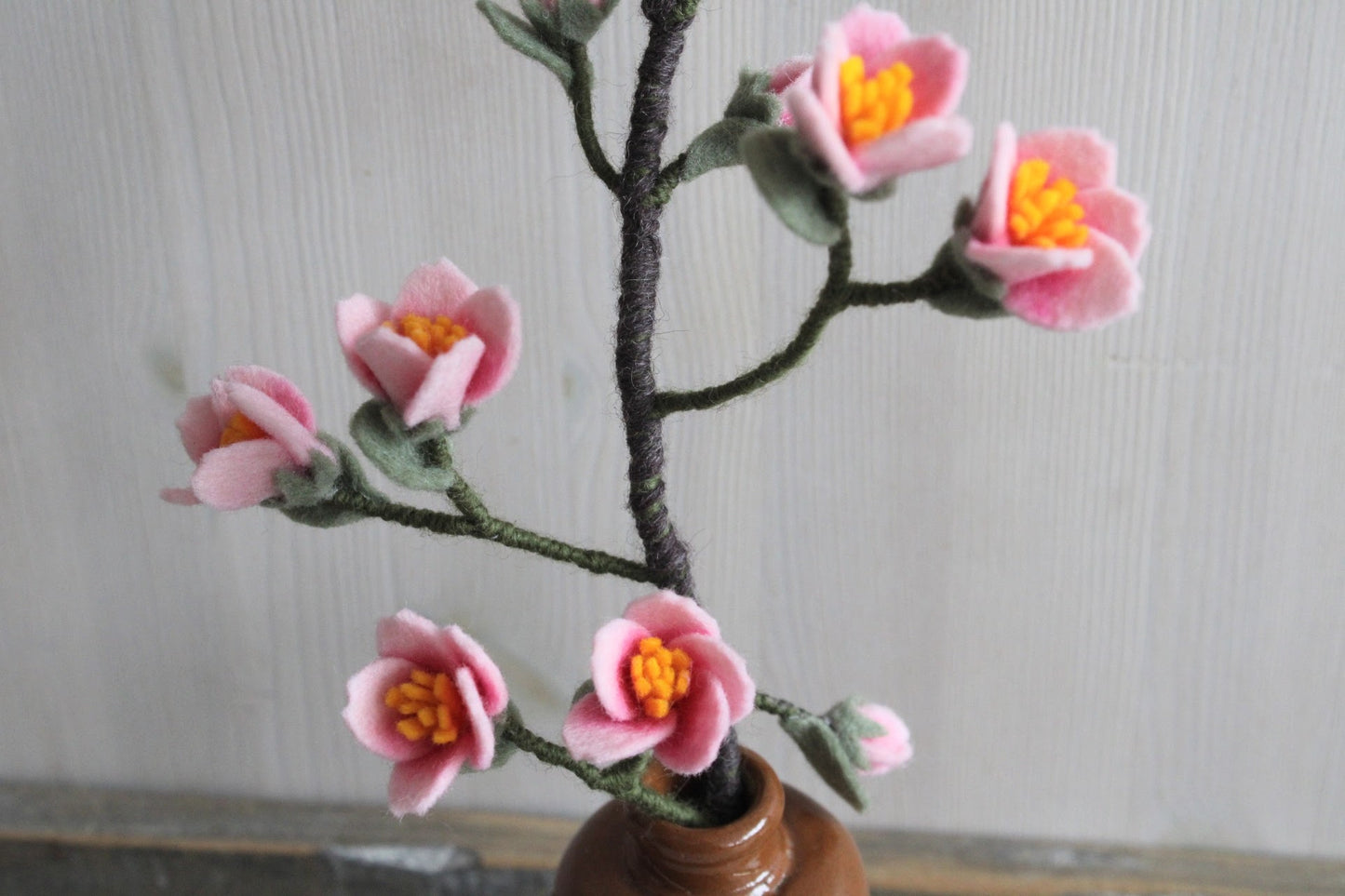 Handgemaakte kersenbloesemtak van vilt met zachte roze bloemetjes en groene blaadjes, geplaatst in een bruine keramieken vaas op een rustieke houten ondergrond. Een tijdloze en duurzame decoratie, perfect voor een lentegevoel in huis.