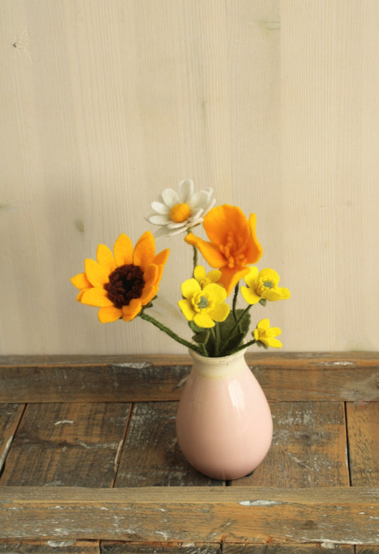 Klein roze vaasje met viltbloemen zonnebloem, boterbloem, klaproos en margriet op een houten ondergrond