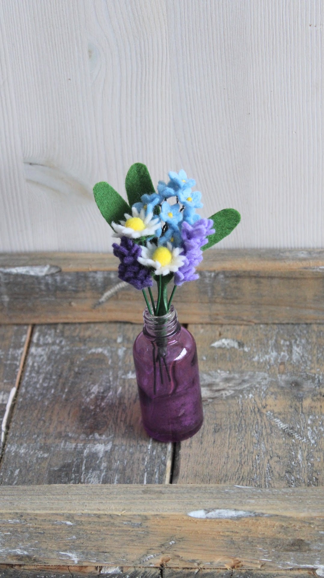 handgemaakt mini boeketje met madeliefjes vergeet-me-nietje en lavendel in een paars glazen vaasje op een houten plateau met een lichte achtergrond