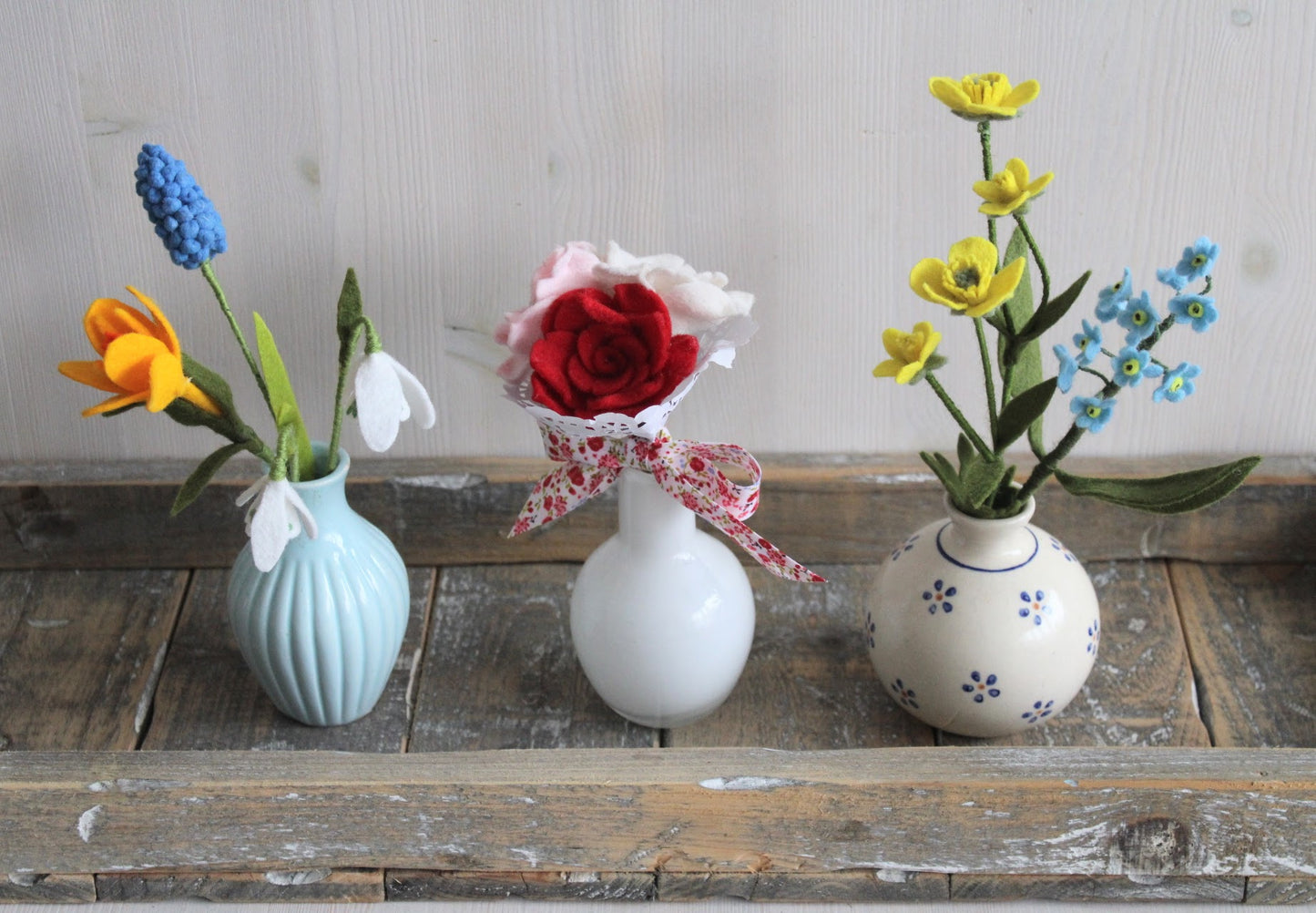 Drie handgemaakte boeketjes van vilt in drie verschillende vaasjes. blauw vaasje met sneeuwklokjes blauw druifje en krokus, wit vaasje met biedermeiertje roosjes en gebloemd vaasje met boterbloem en vergeet-me-nietjes op een houten ondergrond