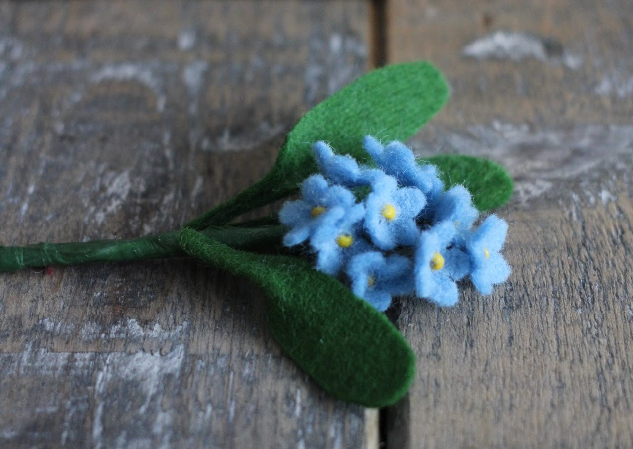 Handgemaakte vilten vergeet me nietje met blauwe bloemetjes en een geel kralenhartje, op een korte steel bekleed met groene wol en drie groene blaadjes. liggend op een rustieke houten ondergrond. Perfect als duurzame decoratie of als onderdeel van een viltbloemenboeketje. het kan ook perfect als broche gemaakt worden
