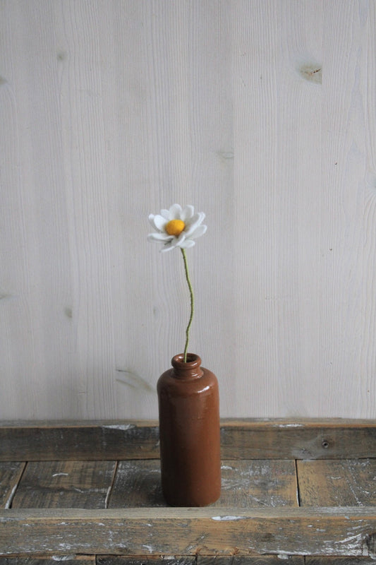 Handgemaakte vilten margriet met witte bloemblaadjes en een geel hart, op een lange steel bekleed met groene wol. Geplaatst in een bruine keramieken vaas op een rustieke houten ondergrond. Perfect als duurzame decoratie of als onderdeel van een viltbloemenboeket