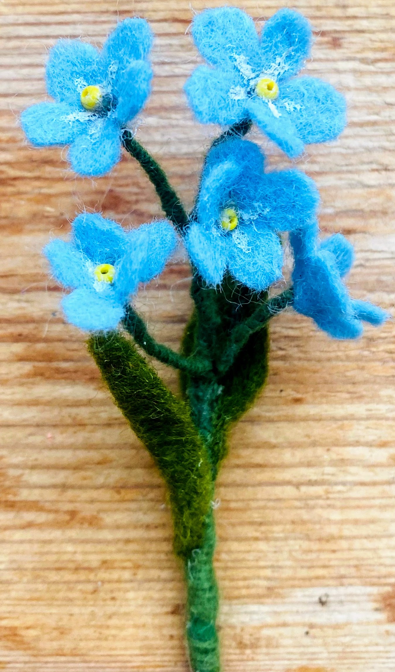Handgemaakte vilten vergeet me nietje met blauwe bloemetjes en een geel kralenhartje, op een korte steel bekleed met groene wol en twee blaadjes. Perfect als duurzame decoratie, als corsage of broche