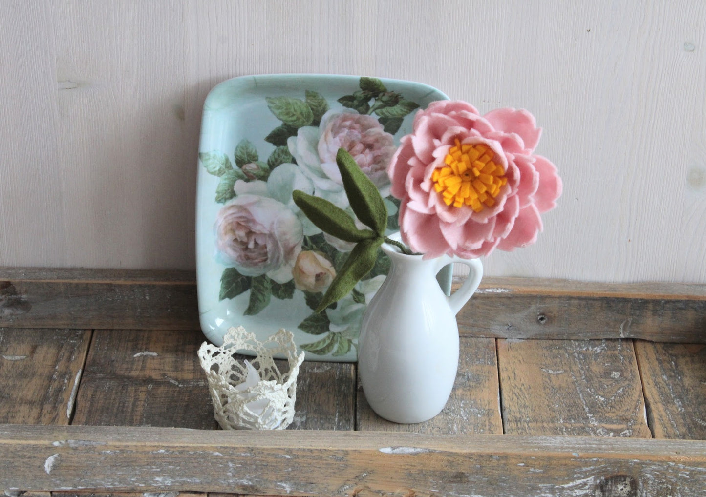 Handgemaakte pioenroos van vilt in een wit keramieken vaasje, met een vintage bloemenbord en een kanten waxinelichthouder op een rustieke houten ondergrond