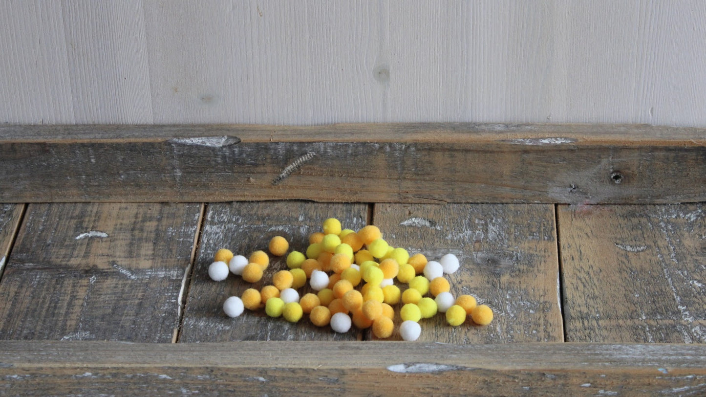 Collectie zachte pompons in de kleuren geel, donkergeel en wit, gebruikt als hartje voor de viltbloemen of voor andere knutselobjecten op een vintage houten oppervlak.