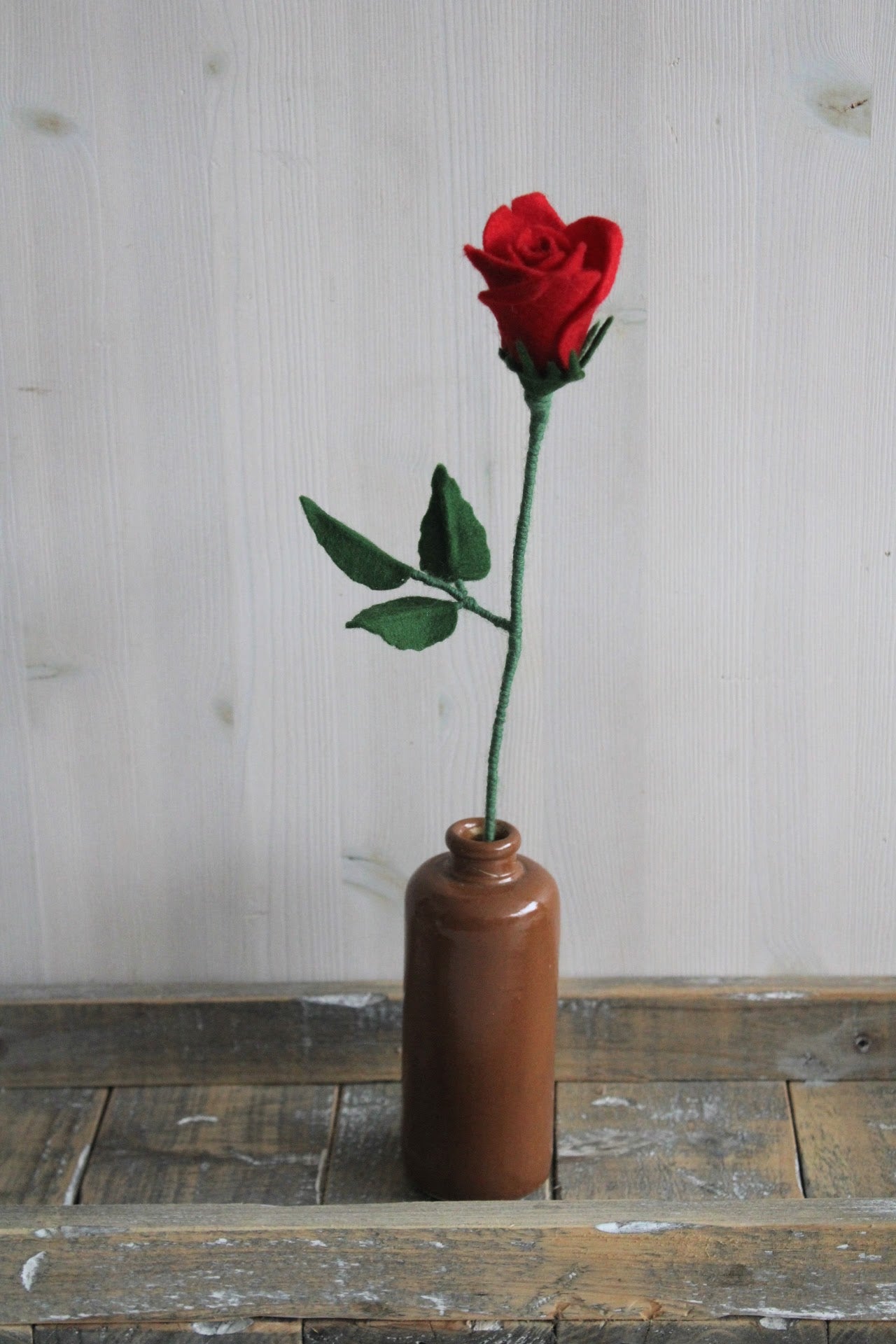 Handgemaakte vilten rode roos op lange steel bekleed met groene wol. Geplaatst in een bruine keramieken vaas op een rustieke houten ondergrond. Perfect als duurzame decoratie of in een blijvend bloeiend viltbloemenboeket