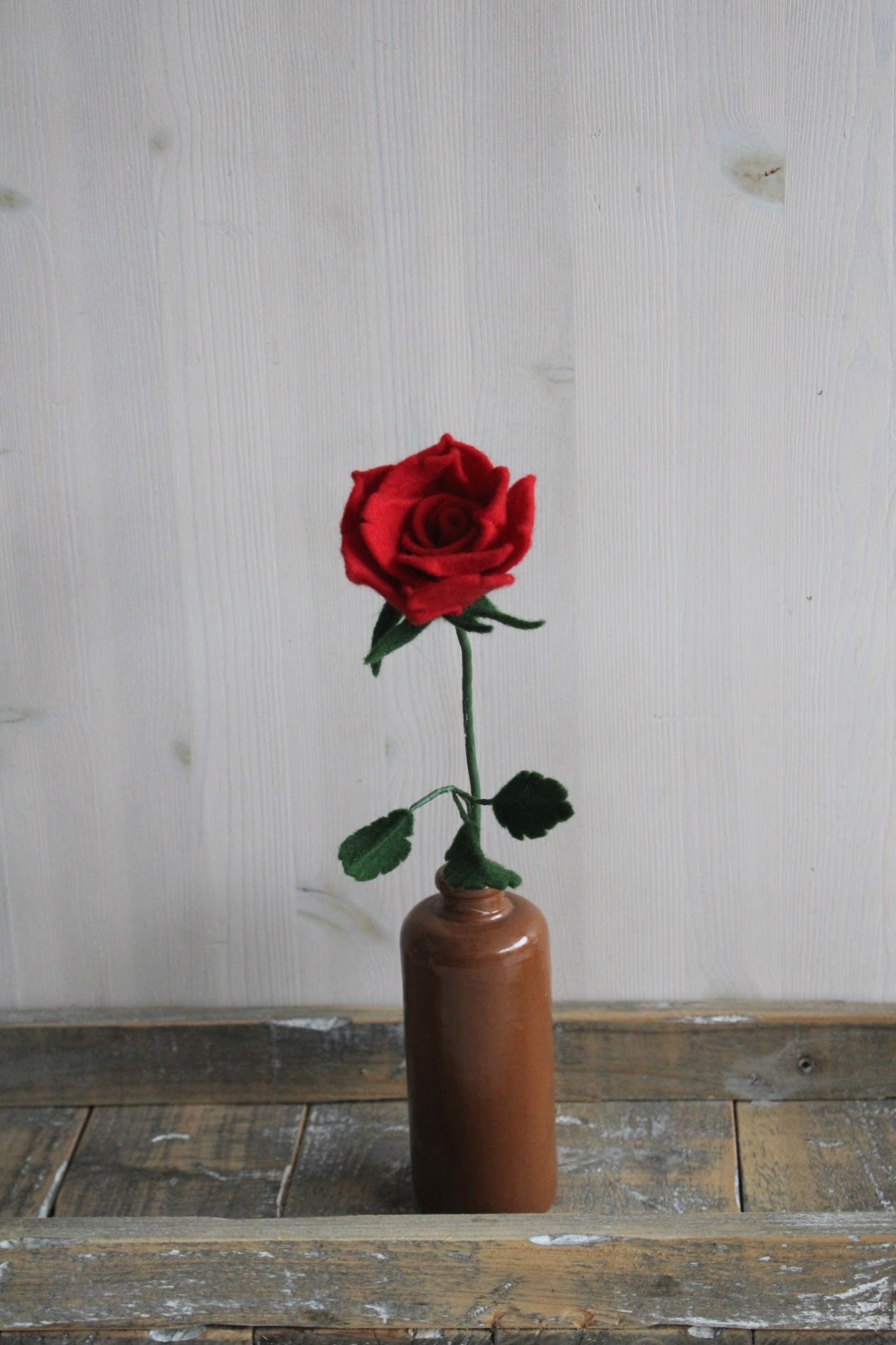 Handgemaakte vilten rode roos op lange steel bekleed met groene wol en blad. Geplaatst in een bruine keramieken vaas op een rustieke houten ondergrond. Perfect als duurzame decoratie of in een blijvend bloeiend viltbloemenboeket