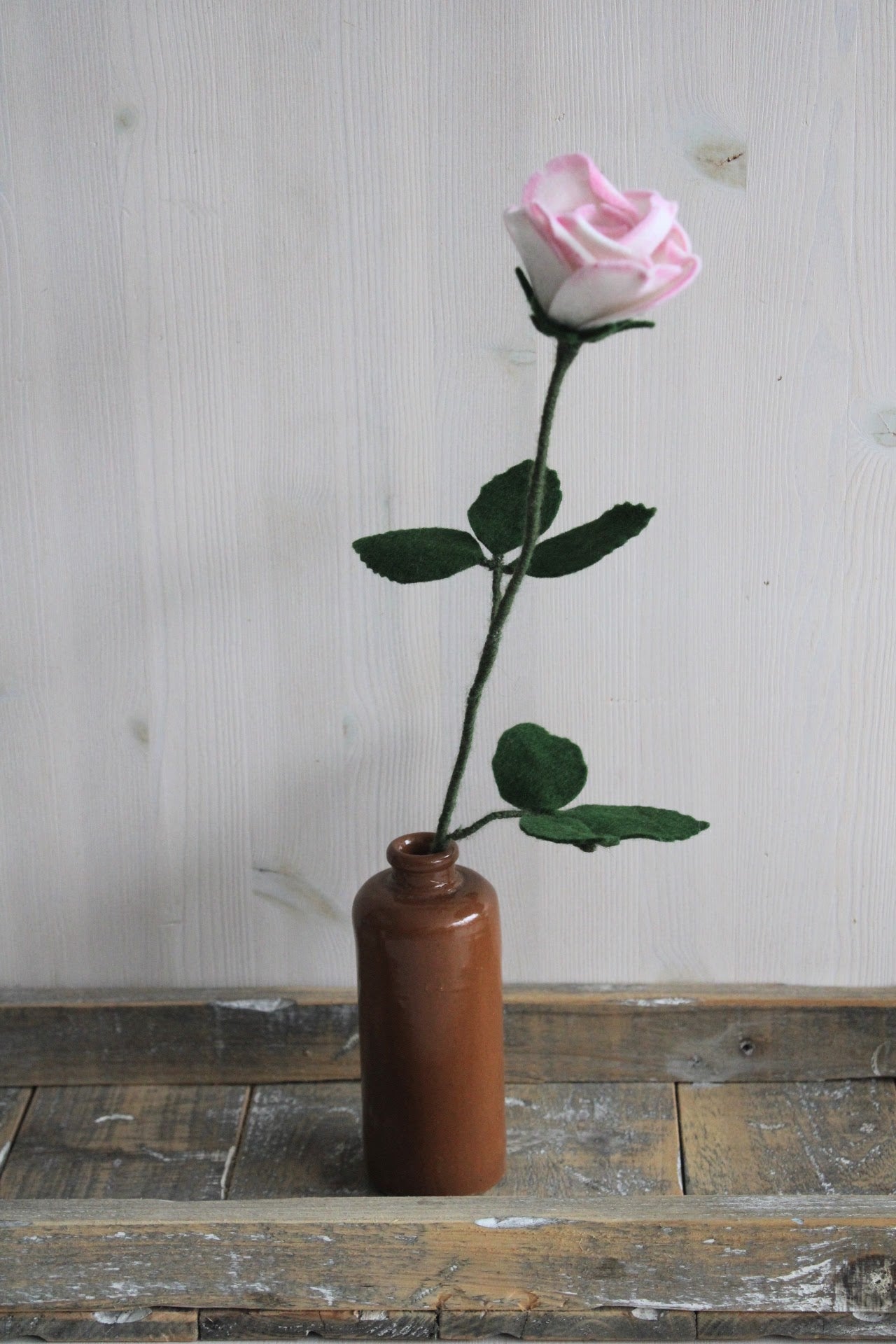 wit met roze roos met bladeren handgemaakt van vilt in een bruine fles op een rustieke houten ondergrond en lichte achtergrond