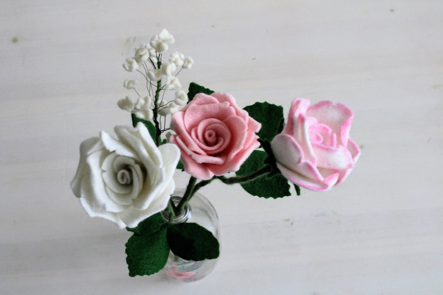 Handgemaakt boeket van viltbloemen in een glazen vaas, met roze en witte rozen en fijn wit gipskruid op een witte ondergrond