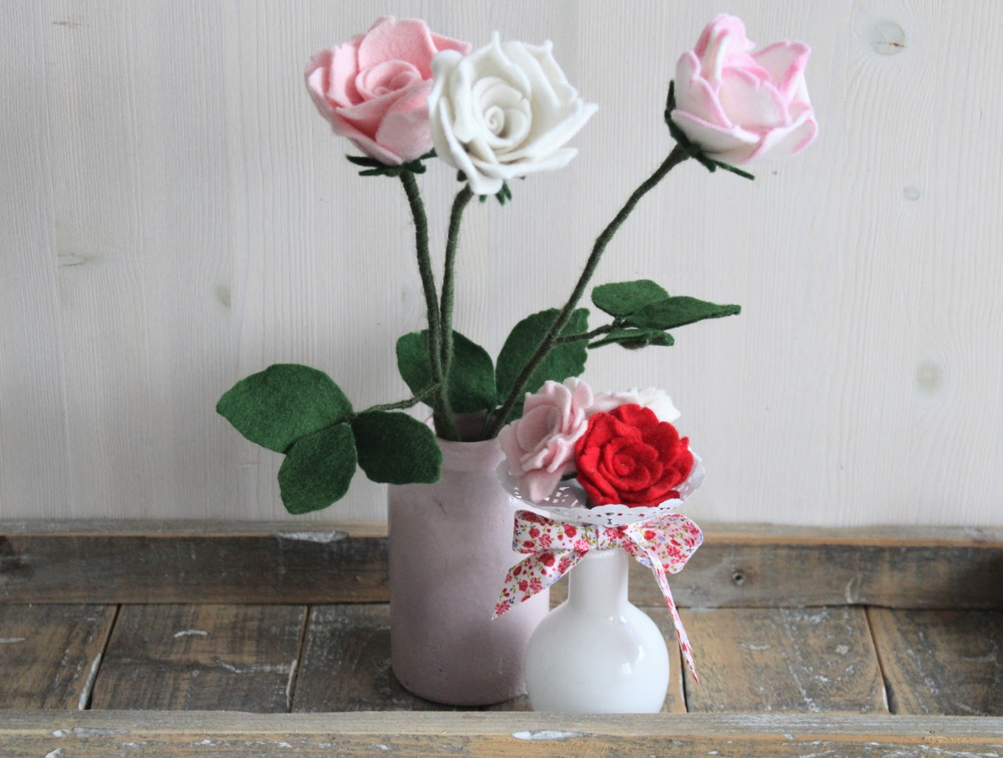 lange vilten Rozen in de kleur roze en wit in een roze vaasje en daarvoor een klein biedermeiertje met drie vilten roosjes rood, wit en roze met gebloemde strik in een wit vaasje op een rustieke houten ondergrond