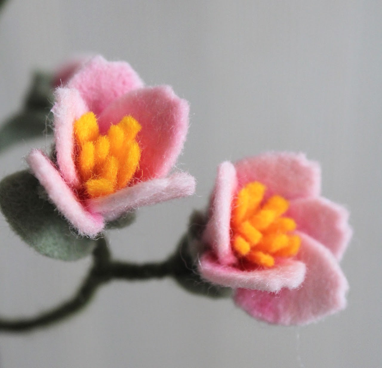 Detailfoto van een handgemaakte vilten kersenbloesem met zachte roze bloemblaadjes, een geel hart en fijne groene details. De stoftextuur van het vilt en ambachtelijke afwerking zijn duidelijk zichtbaar, perfect voor een realistische en duurzame bloemencreatie.