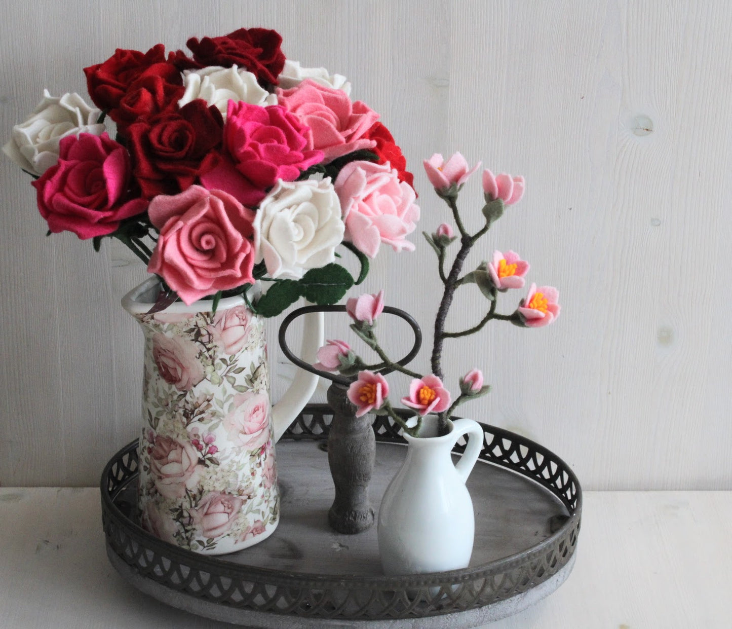 Handgemaakte vilten kersenbloesem in een wit vaasje en een bos verschillende kleuren vilten rozen in een gebloemde vaas op een houten dienblad