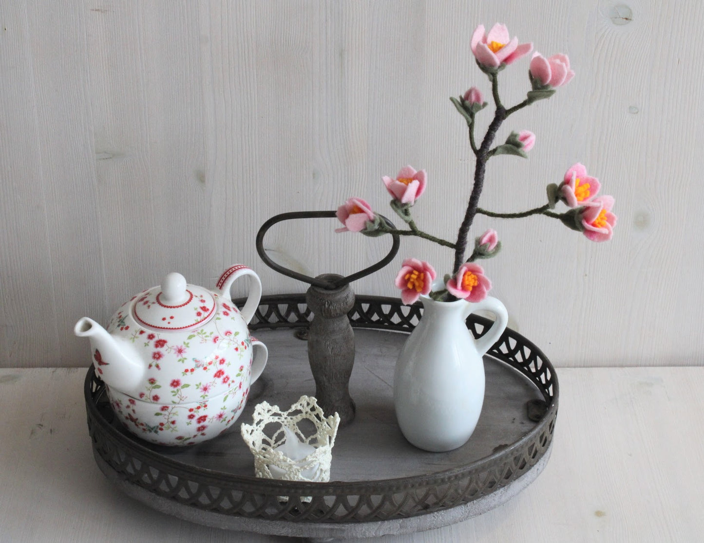 Handgemaakte kersenbloesem van vilt in een wit vaasje met een kanten waxinelichtje en een gebloemde theepot op een houten dienblad