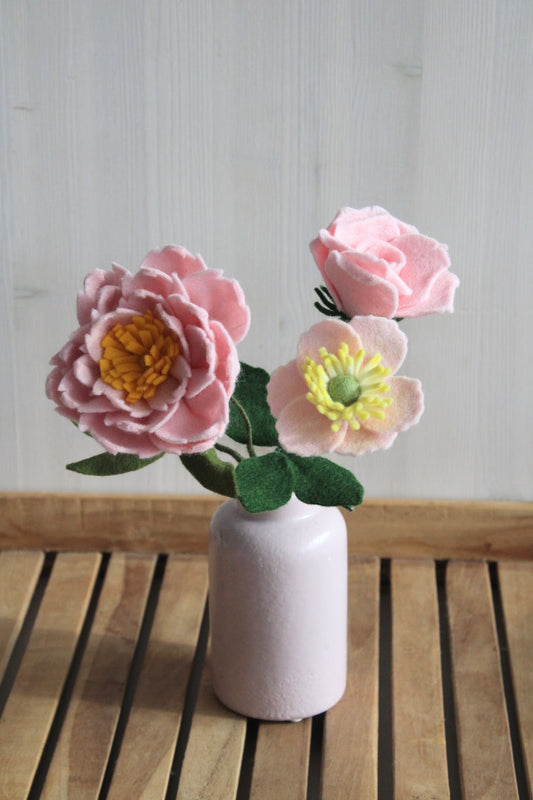 Drie handgemaakte viltbloemen, roze pioenroos, roze anemoon en roze roos in een roze vaasje op een houten ondergrond